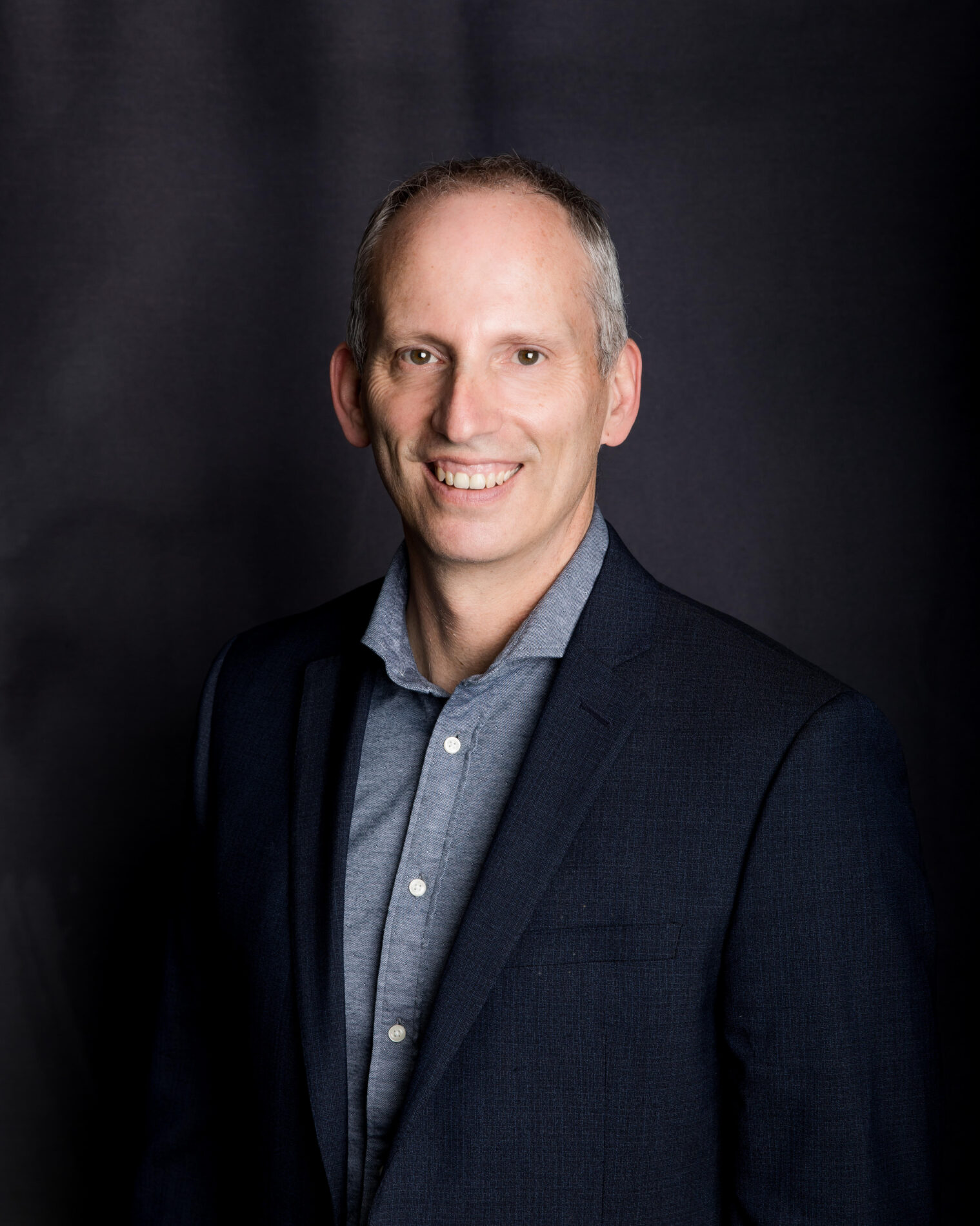 Dale Kuzyk A man in a dark suit and light shirt smiles at the camera against a plain dark background.