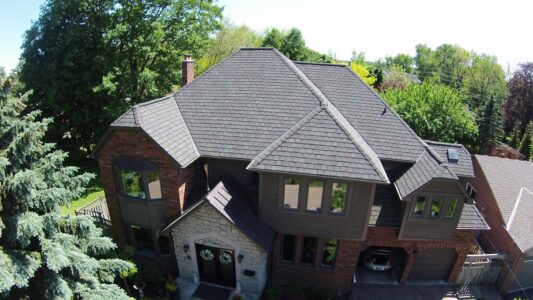 This service is ideal for steep-pitch roofs that cannot be safely climbed, as well as taller buildings without direct roof access. A two-story brick house with gray shingles, multiple rooflines, and a driveway surrounded by trees.